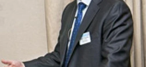 A person wearing a black suit and glasses, standing with their hand on their shoulder. Formal attire and eyewear for polished style.